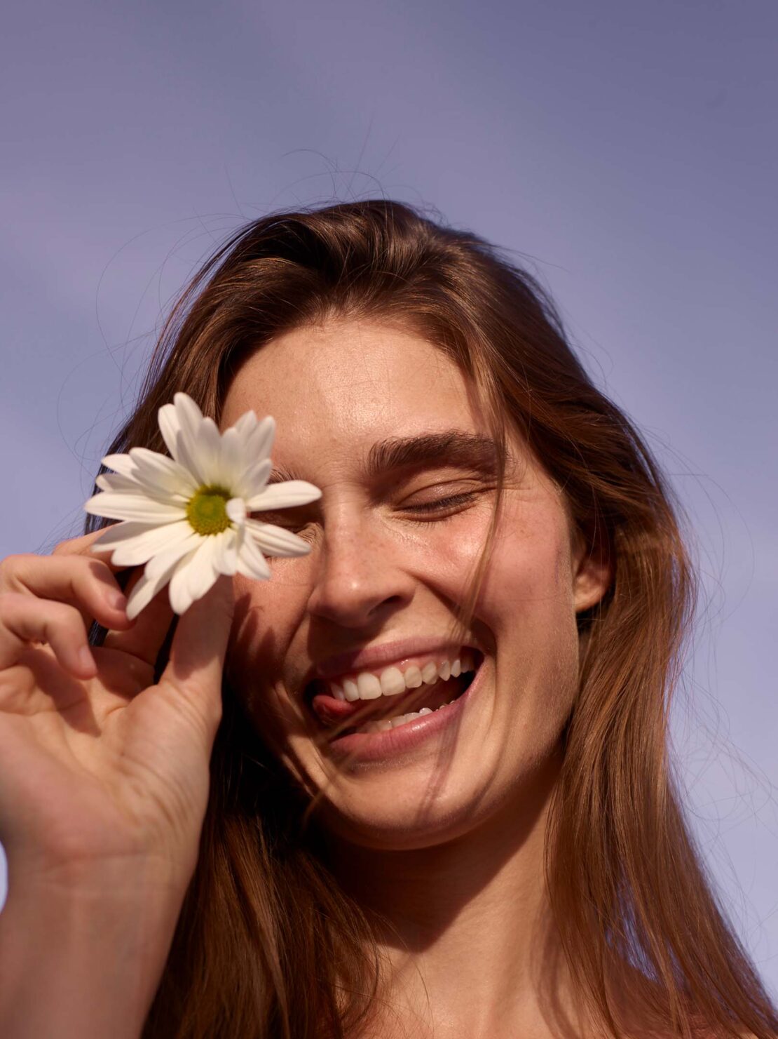 Close-up of a cheerful woman with long brown hair, enjoying the sun with a daisy flower near her eye against a bright sky.