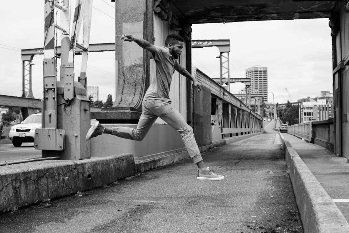 A man is jumping across a concrete divide on a bridge, in a black-and-white urban setting, with city buildings and a vehicle visible in the background.