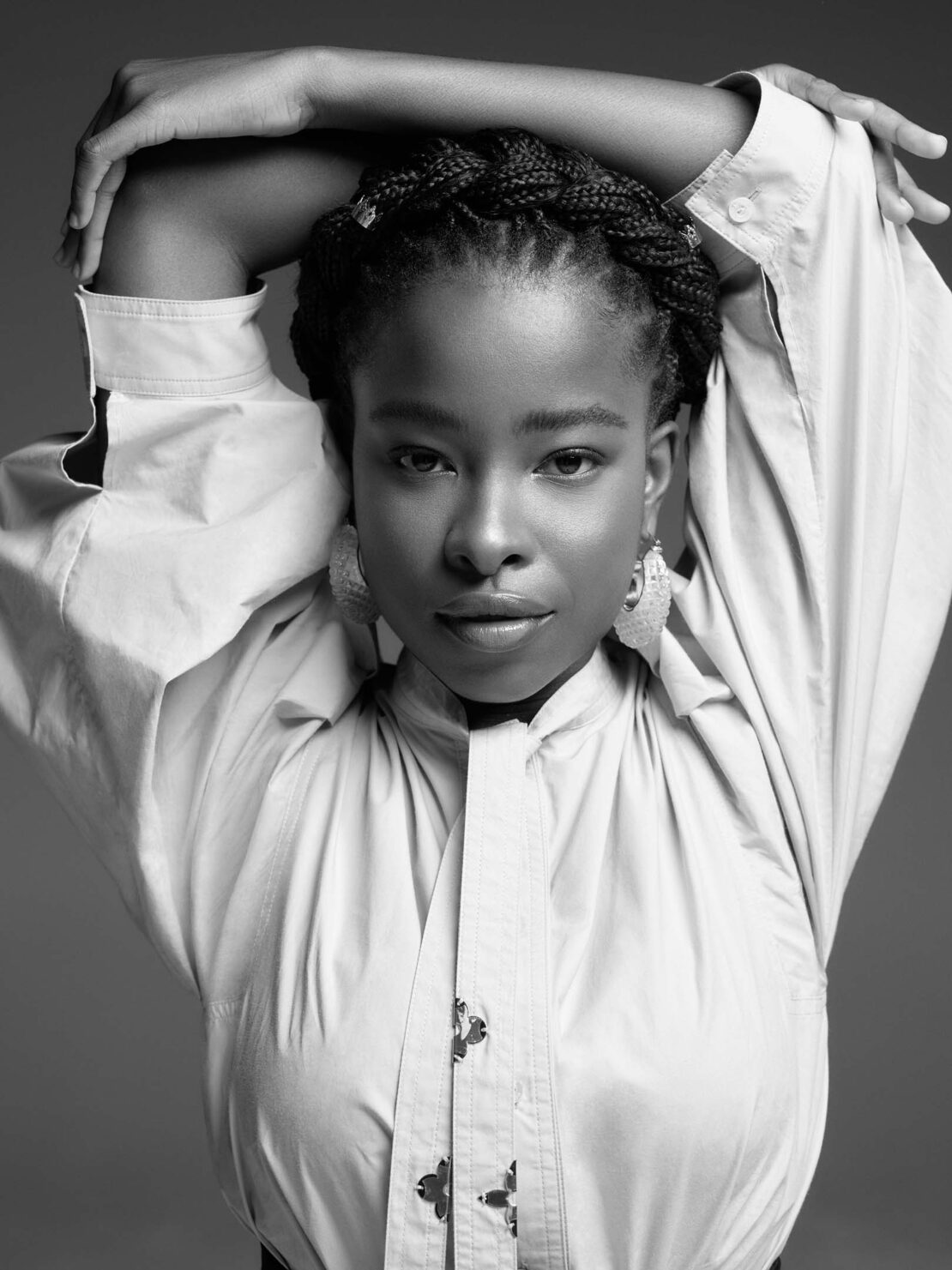 Black and white portrait of a woman with braided hair wearing an oversized button-down shirt, hands crossed behind her head.