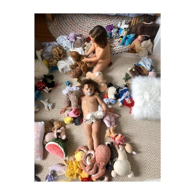 Overhead shot of two young children surrounded by stuffed animals on a patterned rug, creating a playful and whimsical scene.