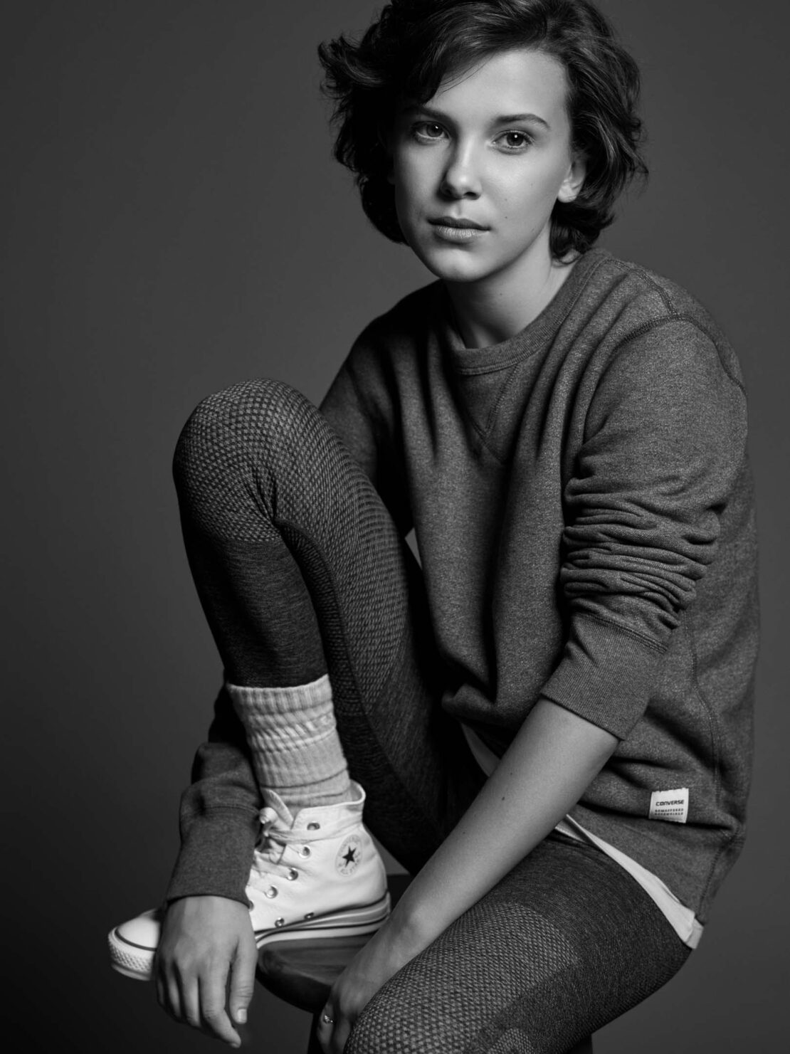 Monochrome portrait of a young woman with short brown hair sitting on a stool wearing a sweatshirt, leggings, socks, and high-top sneakers.