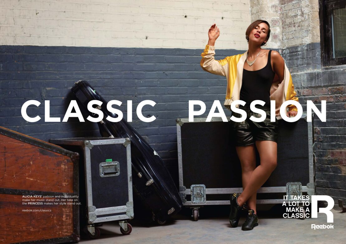 Alicia Keys poses indoors wearing black tank top and yellow jacket, next to music equipment cases, advertising Reebok Classics line.