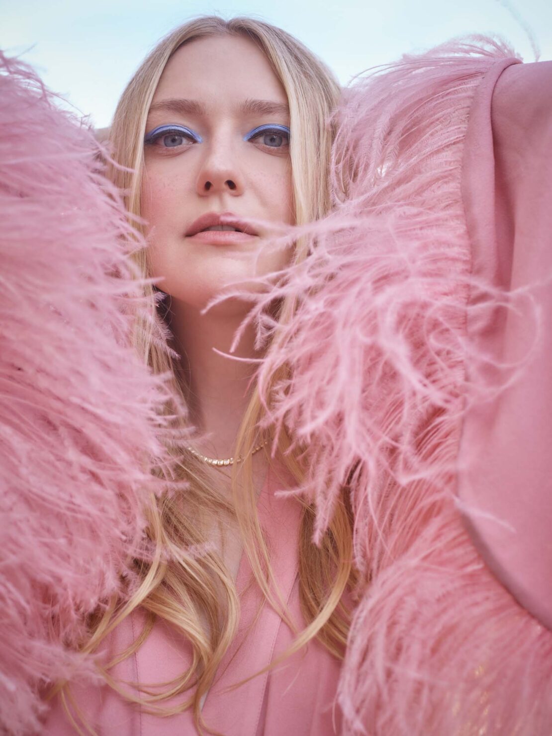 Close-up portrait of a blonde woman with blue eyeshadow, wearing a pink feathered jacket against a soft blue sky. Beauty and fashion.