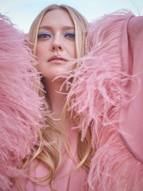 Close-up portrait of a blonde woman with blue eyeshadow, wearing a pink feathered jacket against a soft blue sky. Beauty and fashion.