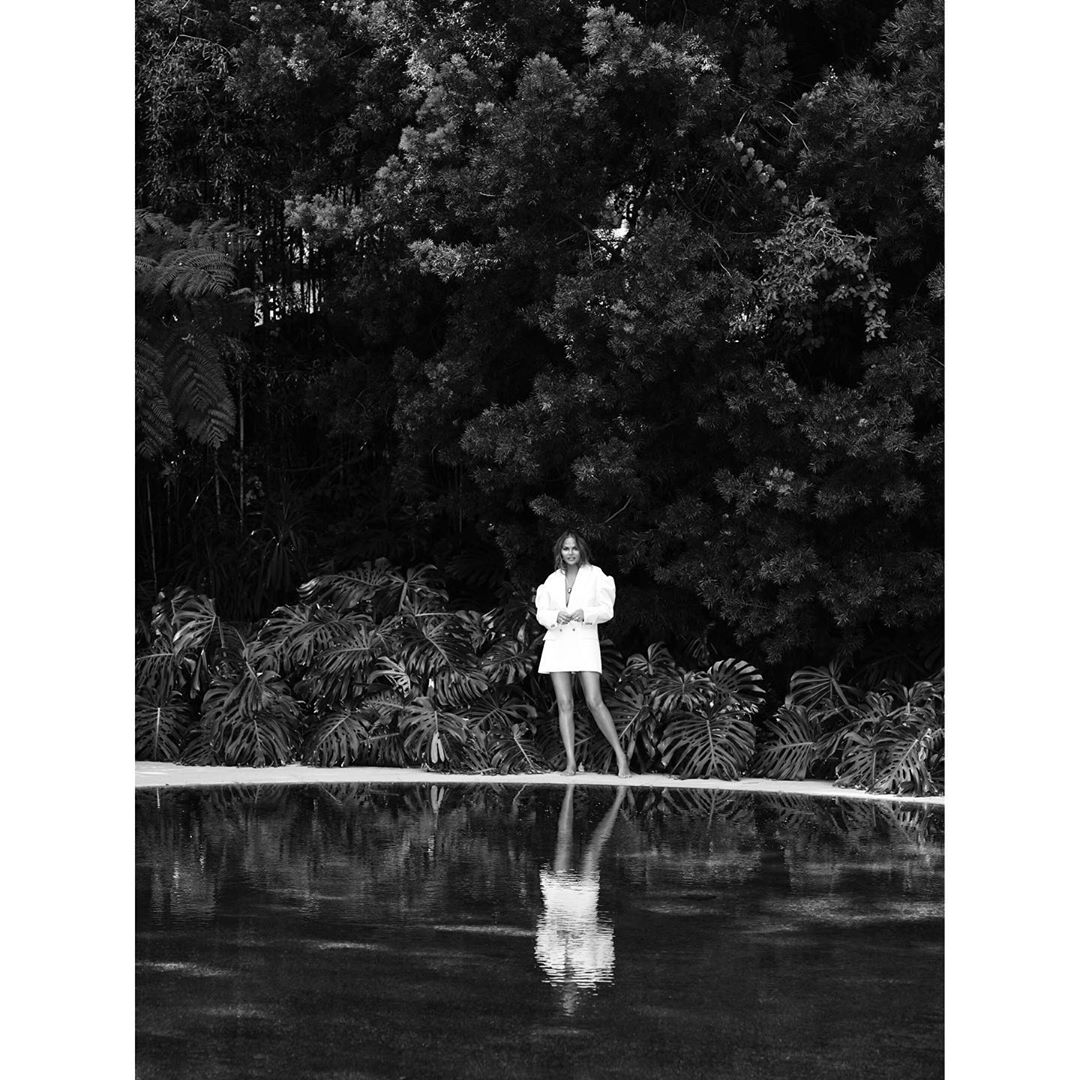 Woman in white blazer and shorts stands in front of reflective water near lush vegetation in black and white.