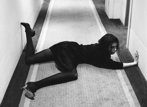 Woman in a black dress and heels lying flat on a hotel hallway floor with her hand against the wall, captured in black and white.
