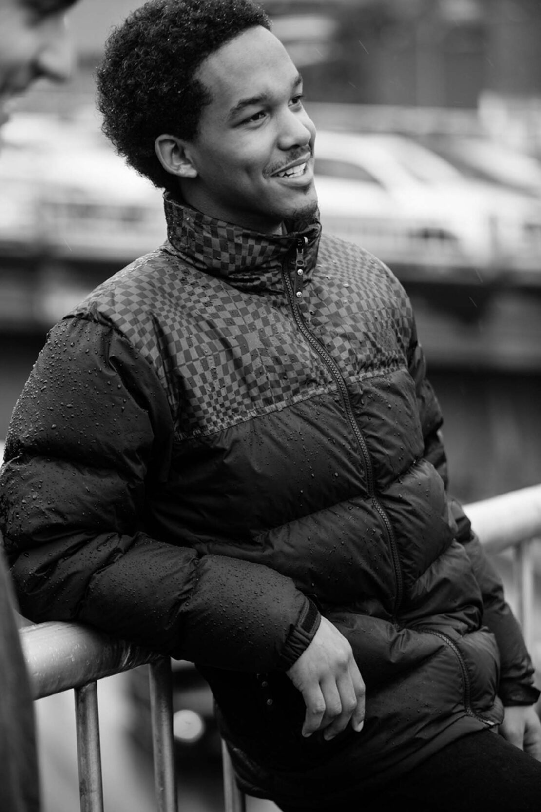 Black and white portrait of a young adult Black male wearing a puffer jacket, leaning on a metal rail, smiling, outdoors on an overcast day.