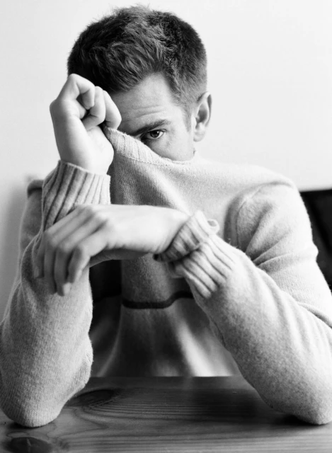 Monochrome portrait of a man with short hair, wearing a sweater, sitting at a table with his face partially obscured by his hand.