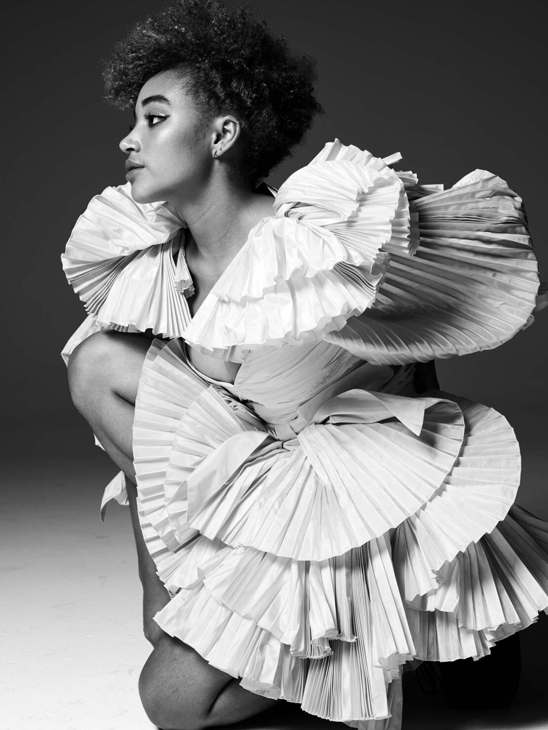 A young woman with natural hair poses kneeling in a studio, wearing a white pleated dress against a grey background in a fashion editorial.