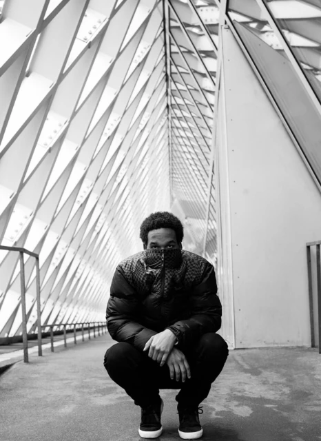 Monochrome portrait of an African-American adult male crouching in a patterned corridor wearing a dark puffer jacket and sneakers.