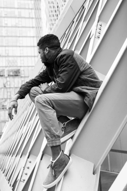 Black and white shot of a Black adult male crouched on a metal structure with a city building in the background, facing left.
