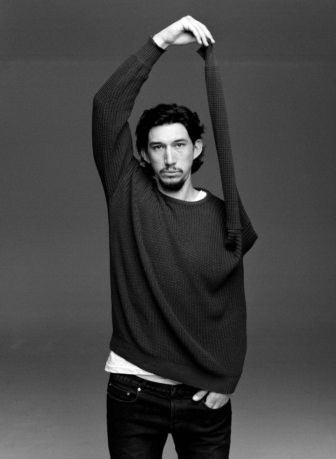 Black and white portrait of Adam Driver in a dark sweater, arms raised, sleeve draped over head, against a neutral gray backdrop.