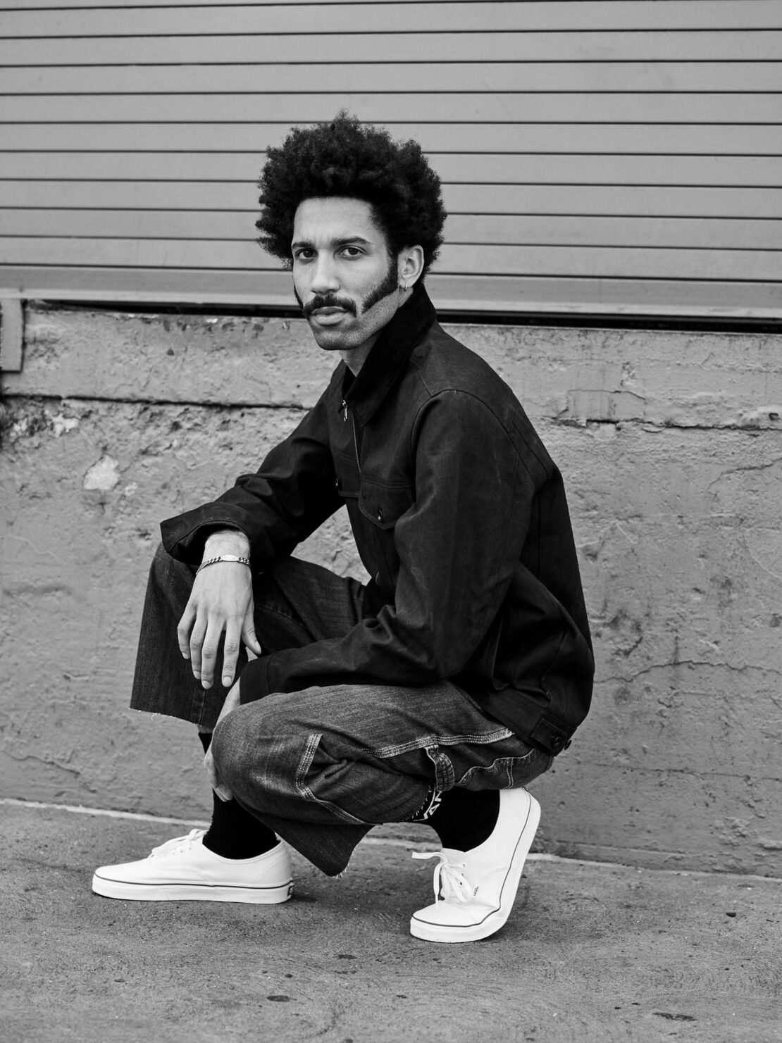 Black and white portrait of a man squatting against an exterior wall wearing a dark jacket, wide trousers, and white sneakers.