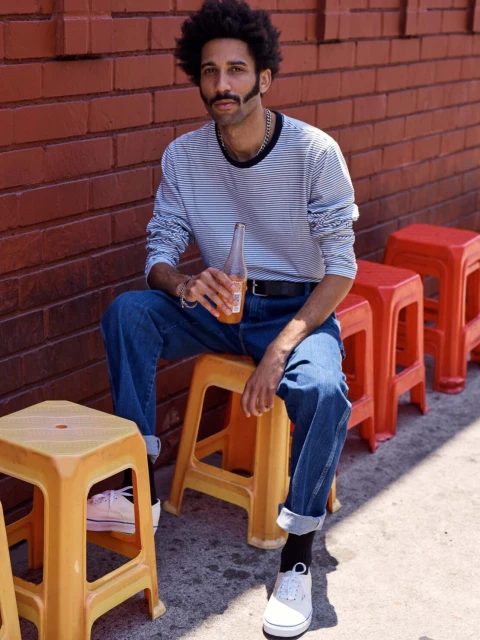 Man in jeans and striped shirt sits on a yellow stool holding a drink, posed in front of a brick wall outdoors on a sunny day.