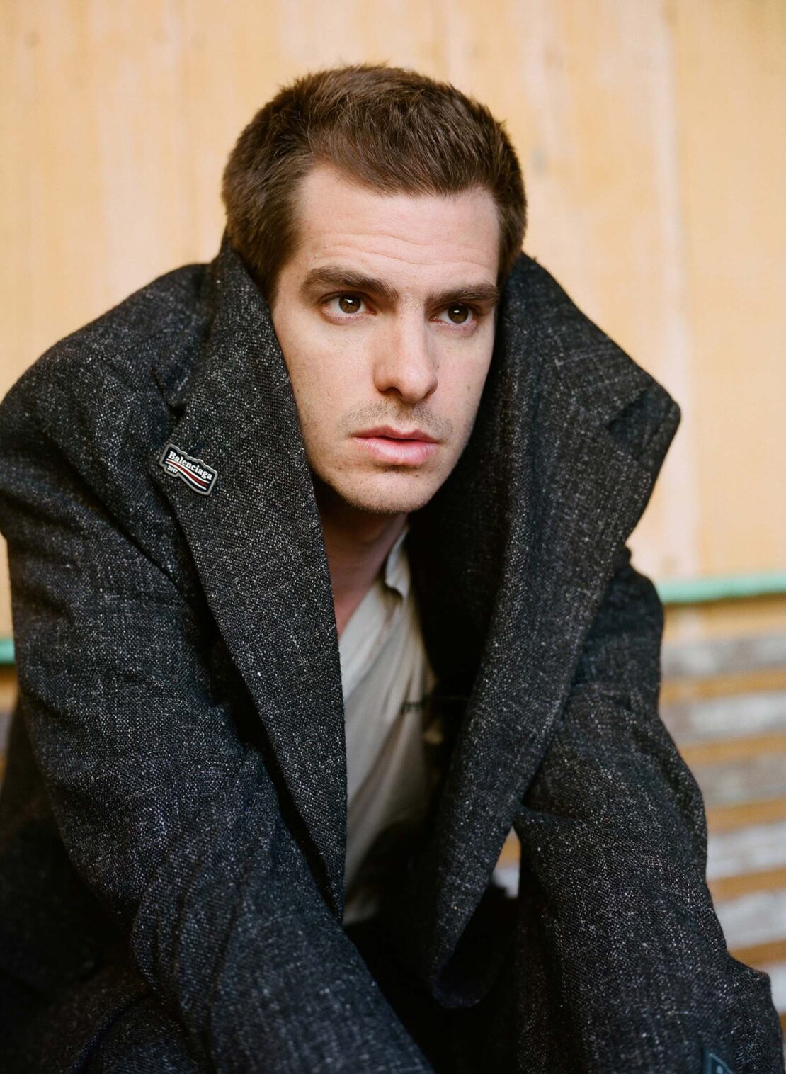 Portrait of Andrew Garfield wearing a textured dark tweed coat with an oversized collar pulled up around his neck.