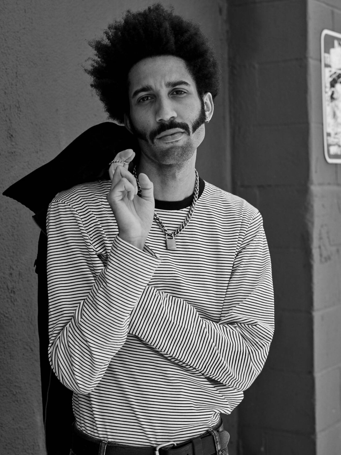 Monochrome portrait of adult male with afro, moustache and sideburns, striped shirt, black jacket over shoulder, against a wall.