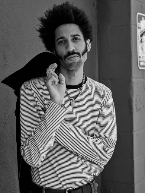 Monochrome portrait of adult male with afro, moustache and sideburns, striped shirt, black jacket over shoulder, against a wall.
