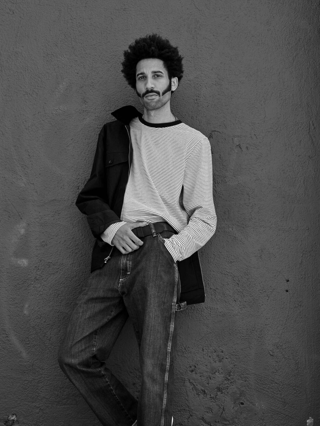 Black and white portrait of a man with an afro hairstyle leaning against a weathered wall, wearing a striped shirt and jeans.