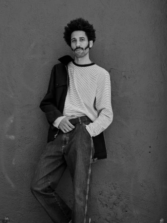 Black and white portrait of a man with an afro hairstyle leaning against a weathered wall, wearing a striped shirt and jeans.