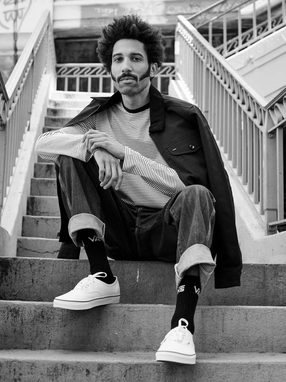 Monochrome portrait of a man with afro and mustache sitting on concrete steps wearing jeans, striped shirt and jacket.