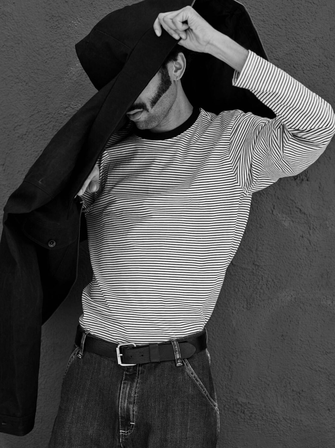 Monochrome portrait of a man in a striped long-sleeve shirt holding a large dark jacket over his head.