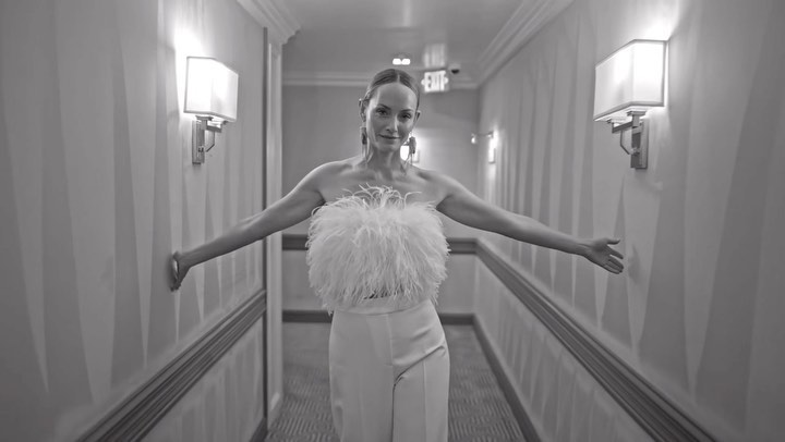Monochrome portrait of a smiling adult woman wearing feather top and wide-leg trousers in a brightly lit hotel corridor.