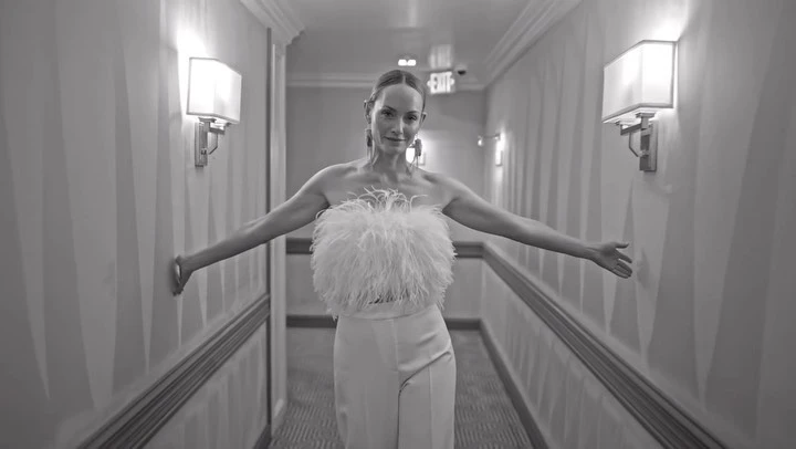 Monochrome portrait of a smiling adult woman wearing feather top and wide-leg trousers in a brightly lit hotel corridor.
