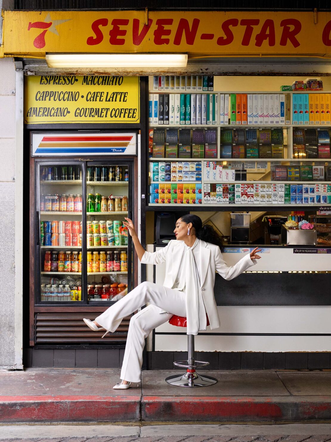 Stylish woman in a white suit poses on a stool outside a Seven-Star convenience store with sodas, snacks, and storefront signage.