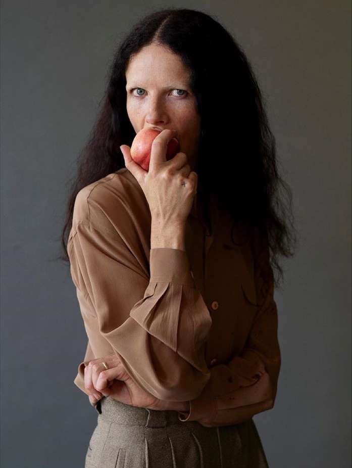 An adult woman with long dark hair and light skin poses indoors eating a red apple, wearing a tan blouse and grey pleated pants.