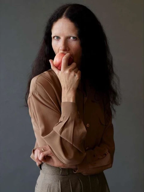 An adult woman with long dark hair and light skin poses indoors eating a red apple, wearing a tan blouse and grey pleated pants.