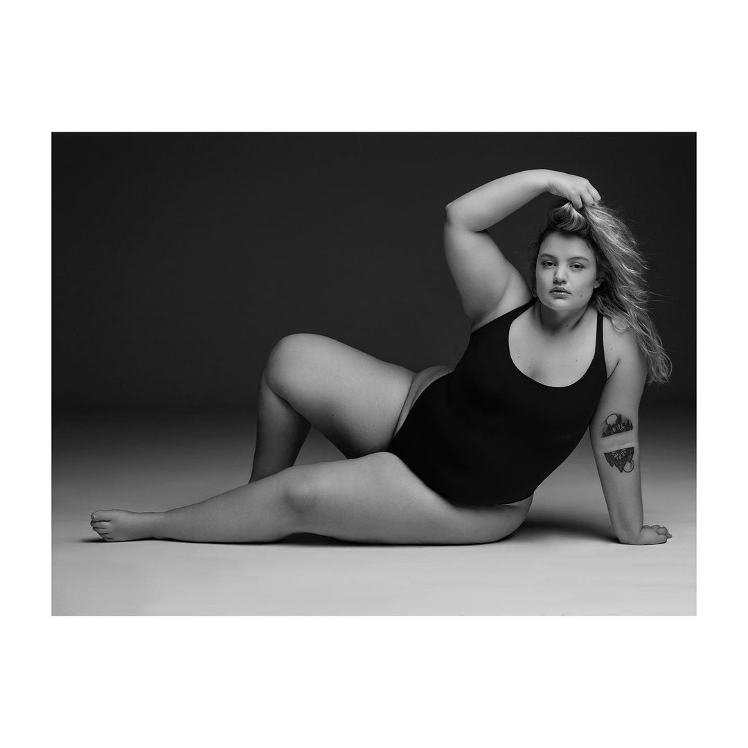 Monochrome studio portrait of a plus-size woman in a black bodysuit, reclining with hand in her hair, gazing at the camera.
