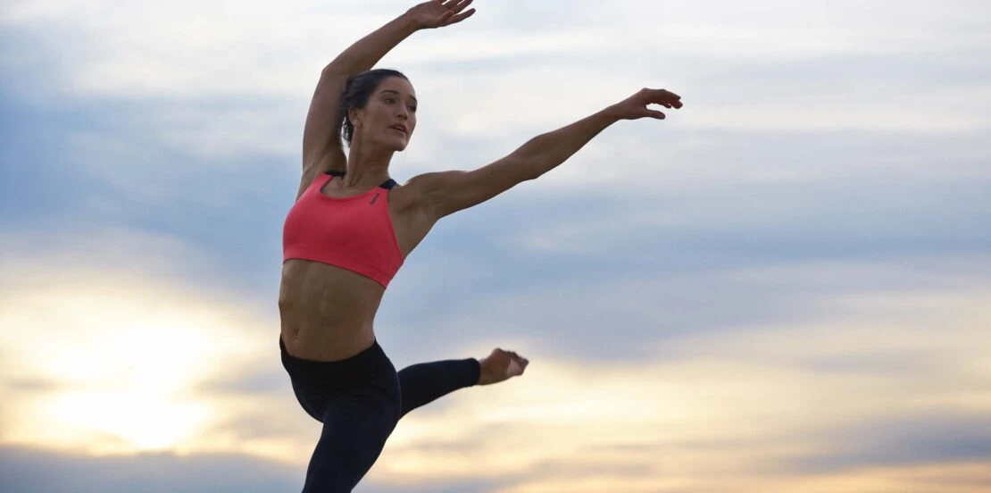 Female athlete mid-jump against a cloudy sky. She wears a sports bra and leggings, with arms extended in a dynamic pose, fitness concept.