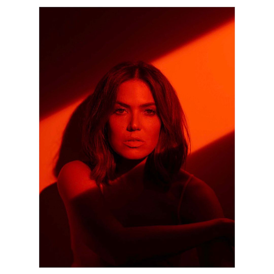 Portrait of a brunette woman posing indoors under warm red lighting with a neutral facial expression and her arms crossed.
