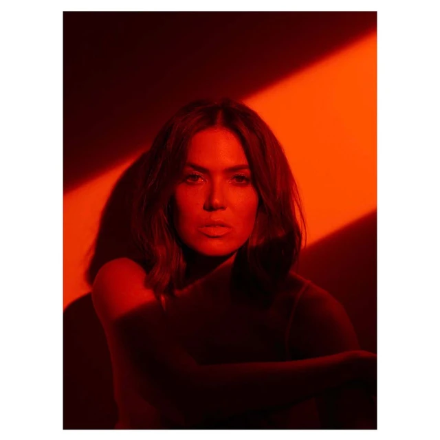 Portrait of a brunette woman posing indoors under warm red lighting with a neutral facial expression and her arms crossed.