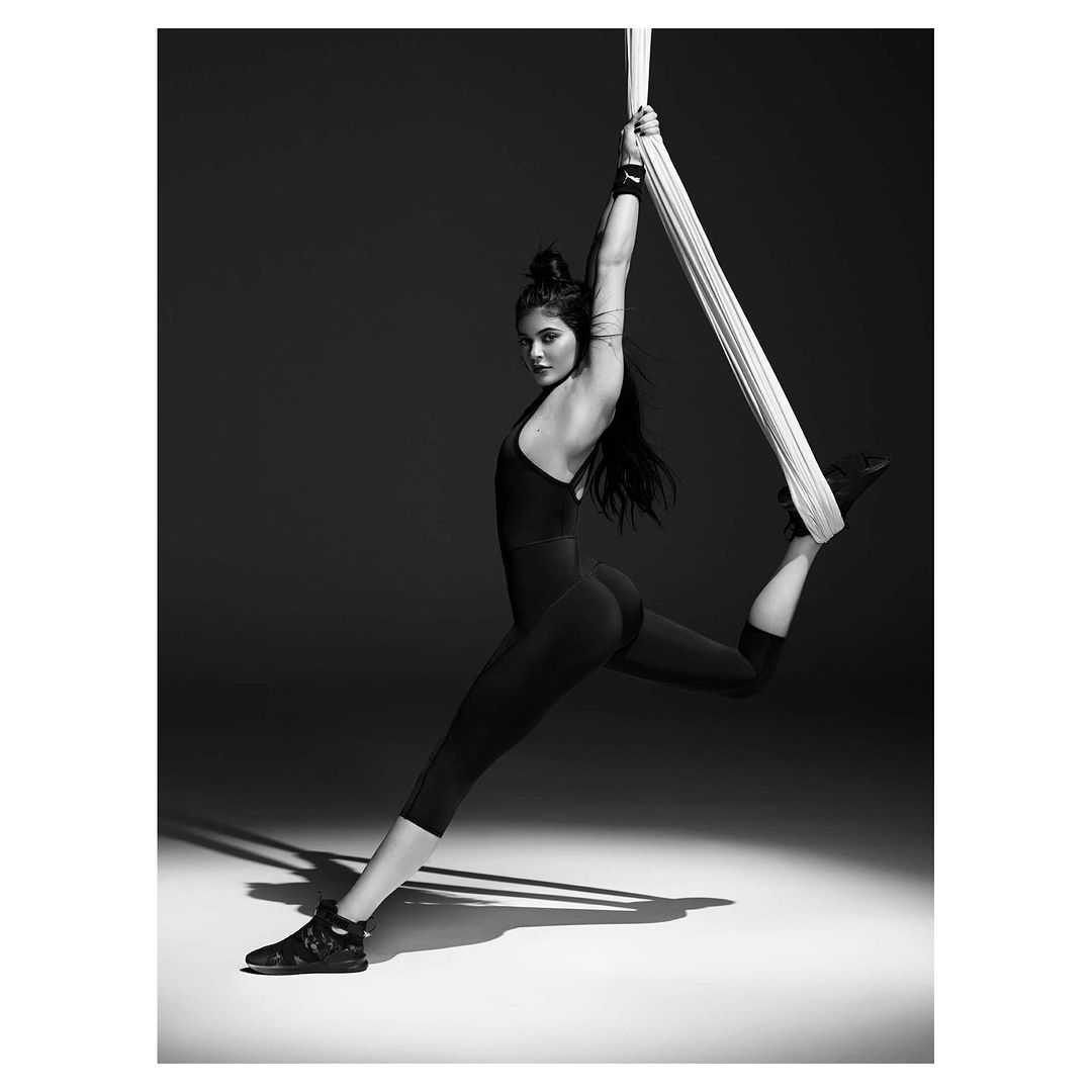 Monochrome portrait of a woman in a black bodysuit suspended mid-air from a white fabric, set against a dark gray background.