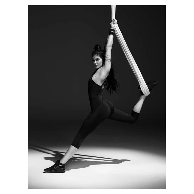 Monochrome portrait of a woman in a black bodysuit suspended mid-air from a white fabric, set against a dark gray background.