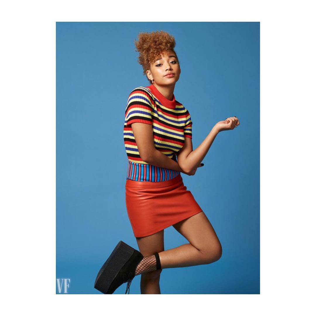 Portrait of a young woman with curly hair wearing a striped top and red skirt, posing against a blue background in a studio.