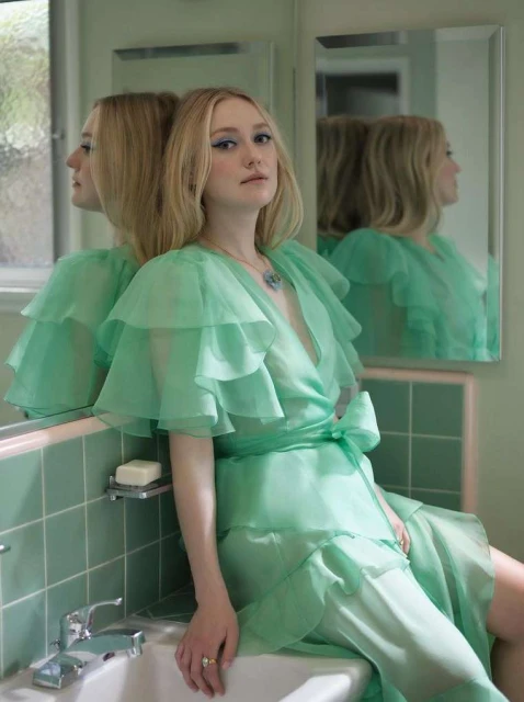 Portrait of adult female in flowing green gown sitting on porcelain sink in bathroom with tile and mirrored wall for fashion editorial.