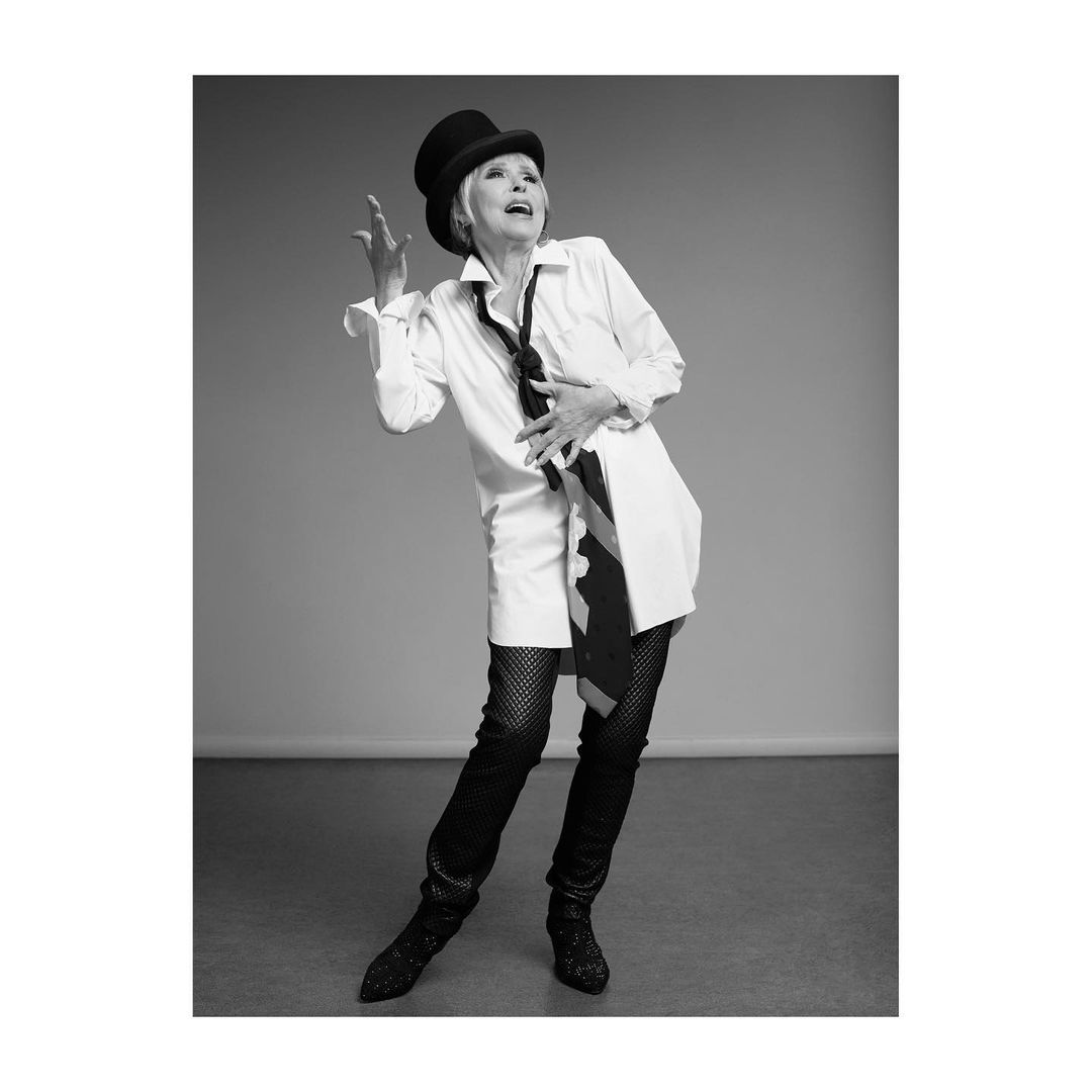 Elegant elderly woman in a top hat, shirt dress, fishnet tights, and high heels poses in a grey photo studio with expressive gestures.