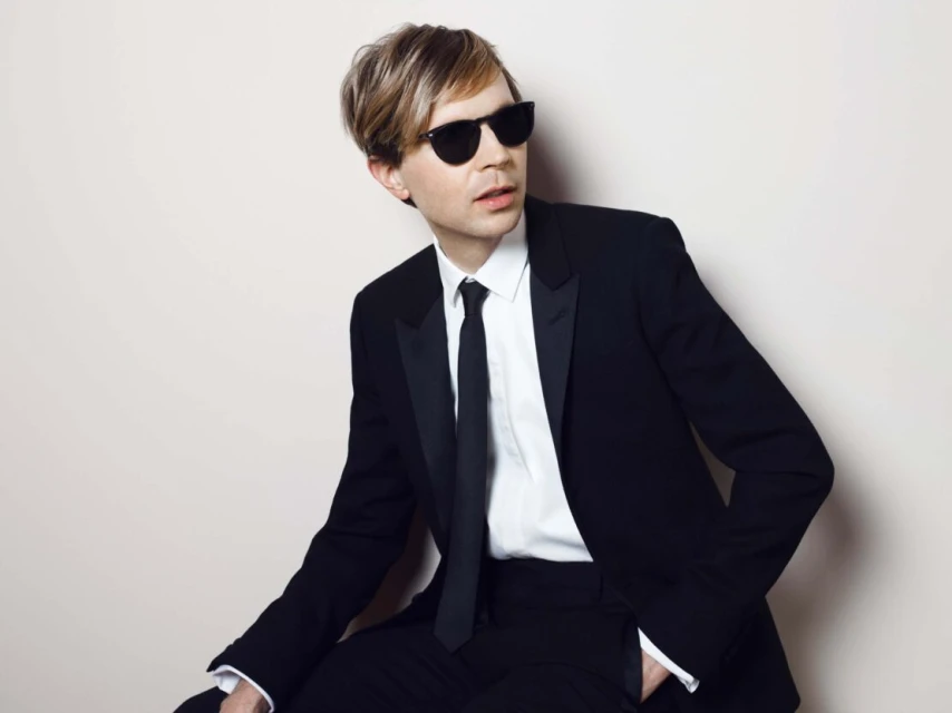 Blond male singer-songwriter in a black tuxedo suit poses indoors wearing sunglasses against a neutral background, looking off-camera.