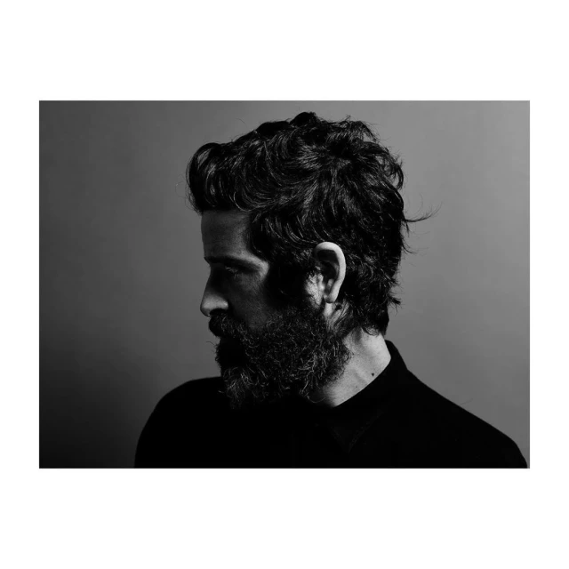 Monochrome profile of a bearded adult male in dark clothing against a gradient grey background, showcasing textured hair and side lighting.