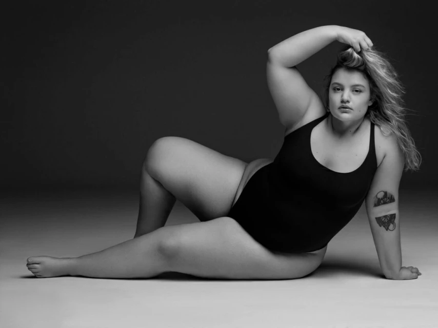 Portrait of a plus-size woman lying on her side wearing a black bodysuit, posing with her hand in her blond hair in a photo studio.
