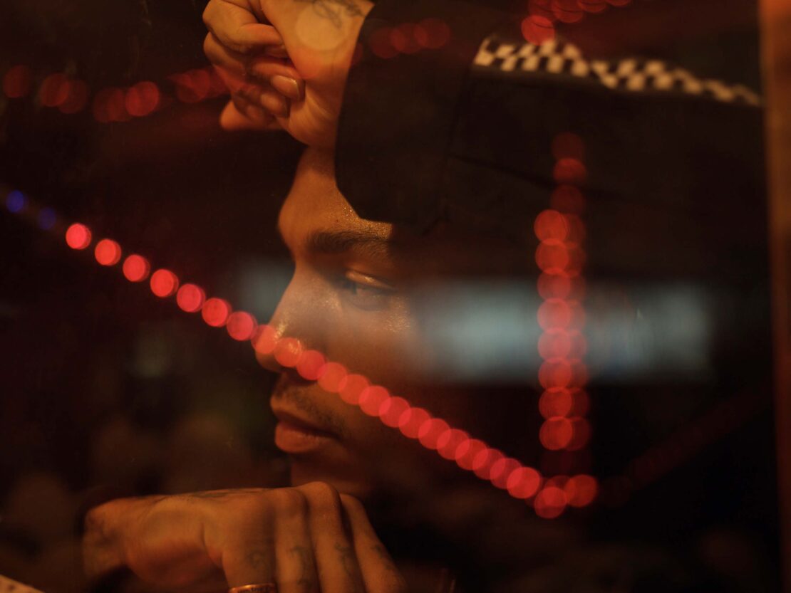 Close-up portrait of a man resting his head on his hands, viewed through a window with glowing red light reflections