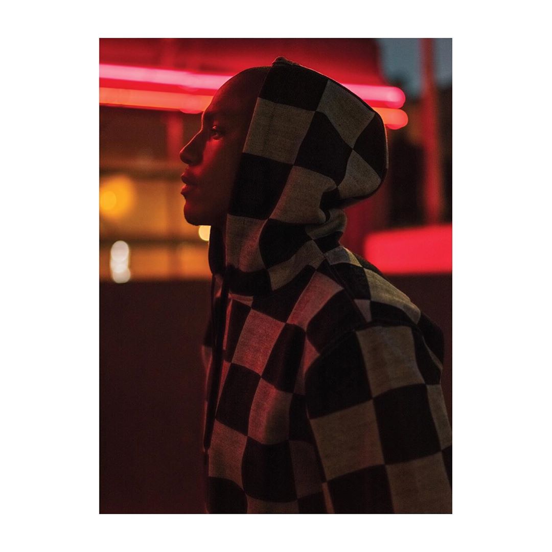 Profile shot of a person in a checkered hooded jacket standing outdoors in a blurred urban environment, illuminated by red neon light.