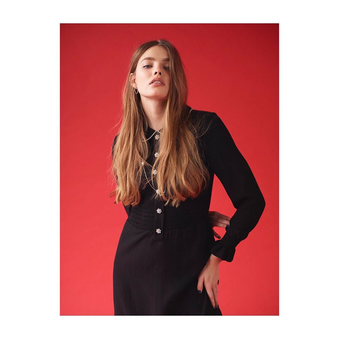 Young woman with long brown hair poses in a long-sleeved black dress with flower-shaped buttons against a red background indoors.