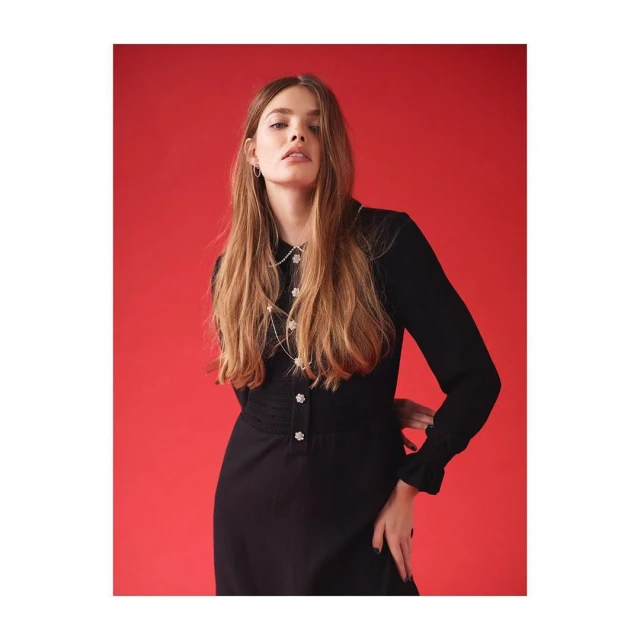 Young woman with long brown hair poses in a long-sleeved black dress with flower-shaped buttons against a red background indoors.