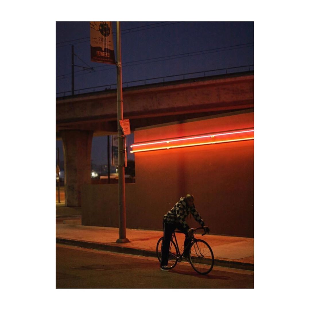 A man rides a bicycle at dusk on a city street with a concrete structure and orange neon lights, creating an urban atmosphere.