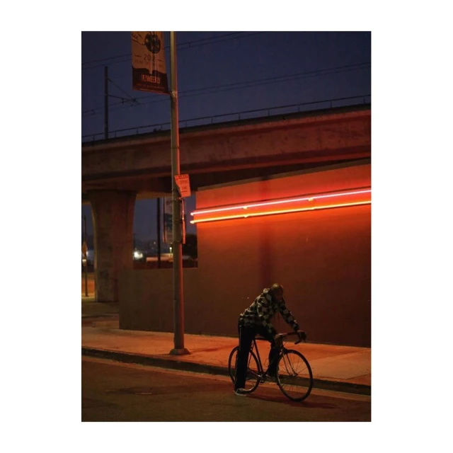 A man rides a bicycle at dusk on a city street with a concrete structure and orange neon lights, creating an urban atmosphere.