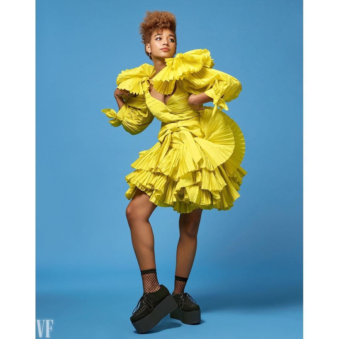 Woman poses in a ruffled yellow dress and platform shoes against a blue background in a fashion forward studio portrait.