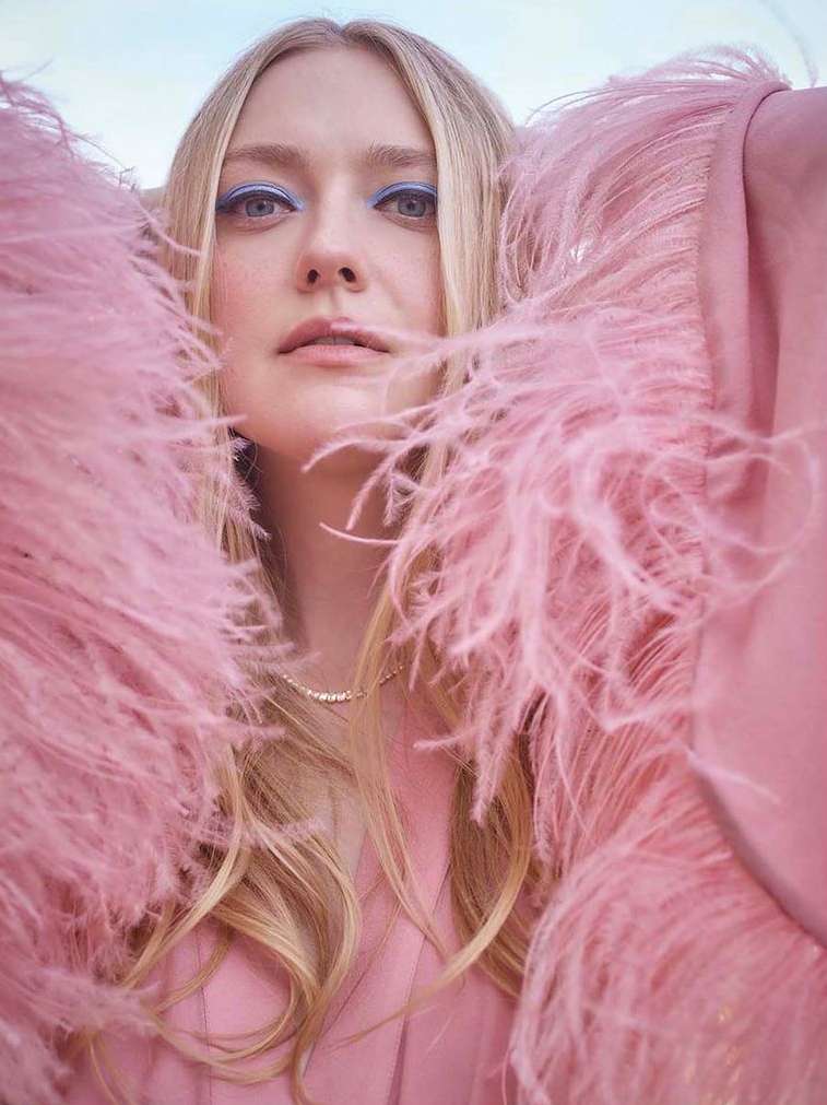 Close-up of blonde woman wearing a pink feathered jacket and blue eye makeup, looking directly at the camera with a neutral expression.